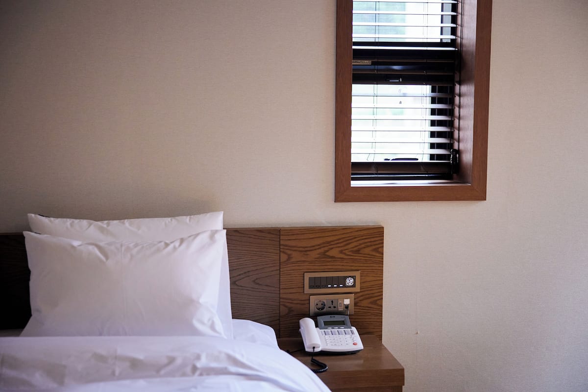A hotel room with a bed, white pillows, a wooden headboard, a bedside telephone, and a window with blinds.