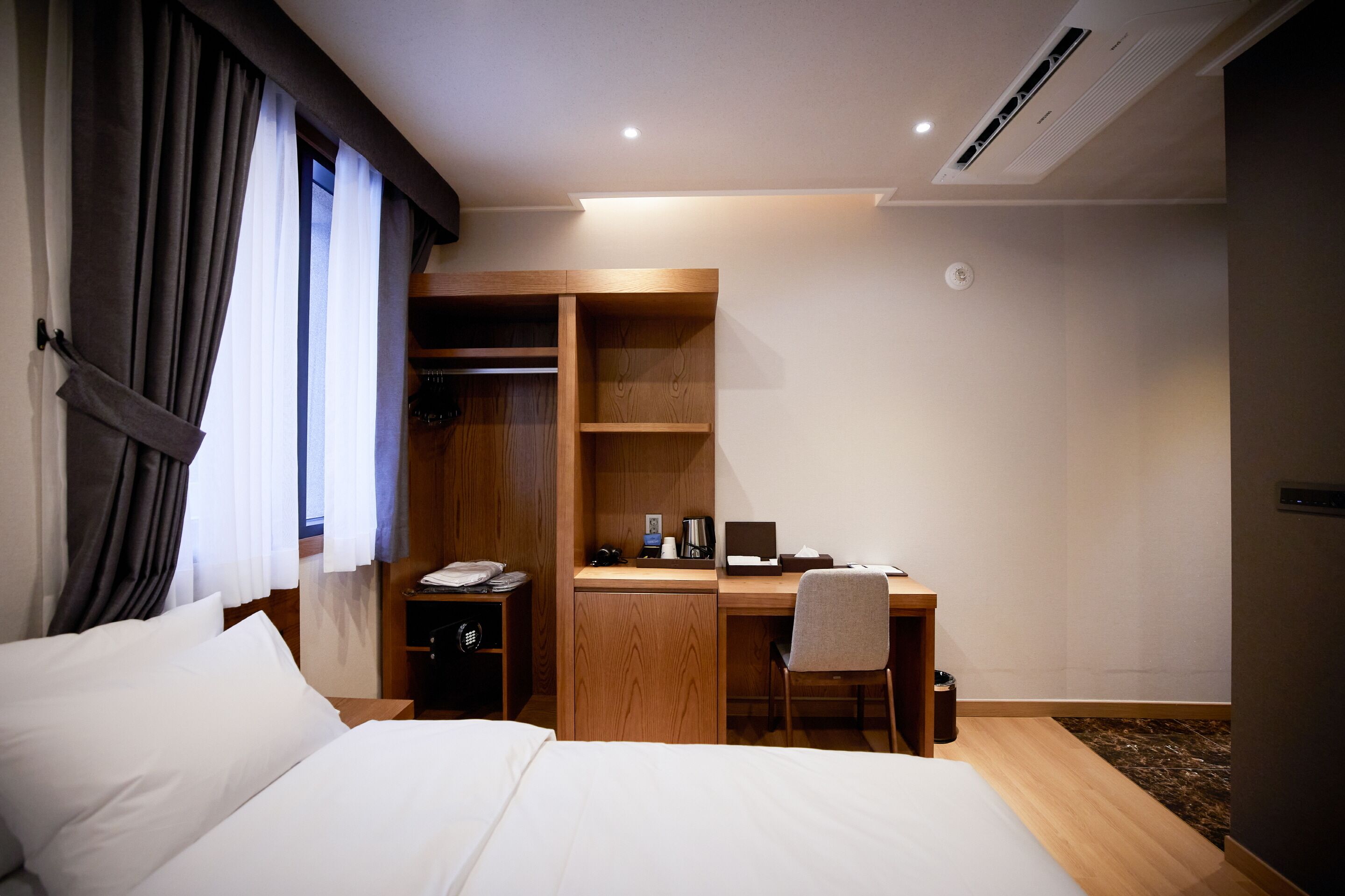 A hotel room with a bed, a desk with a chair, a wardrobe, and a bedside table.