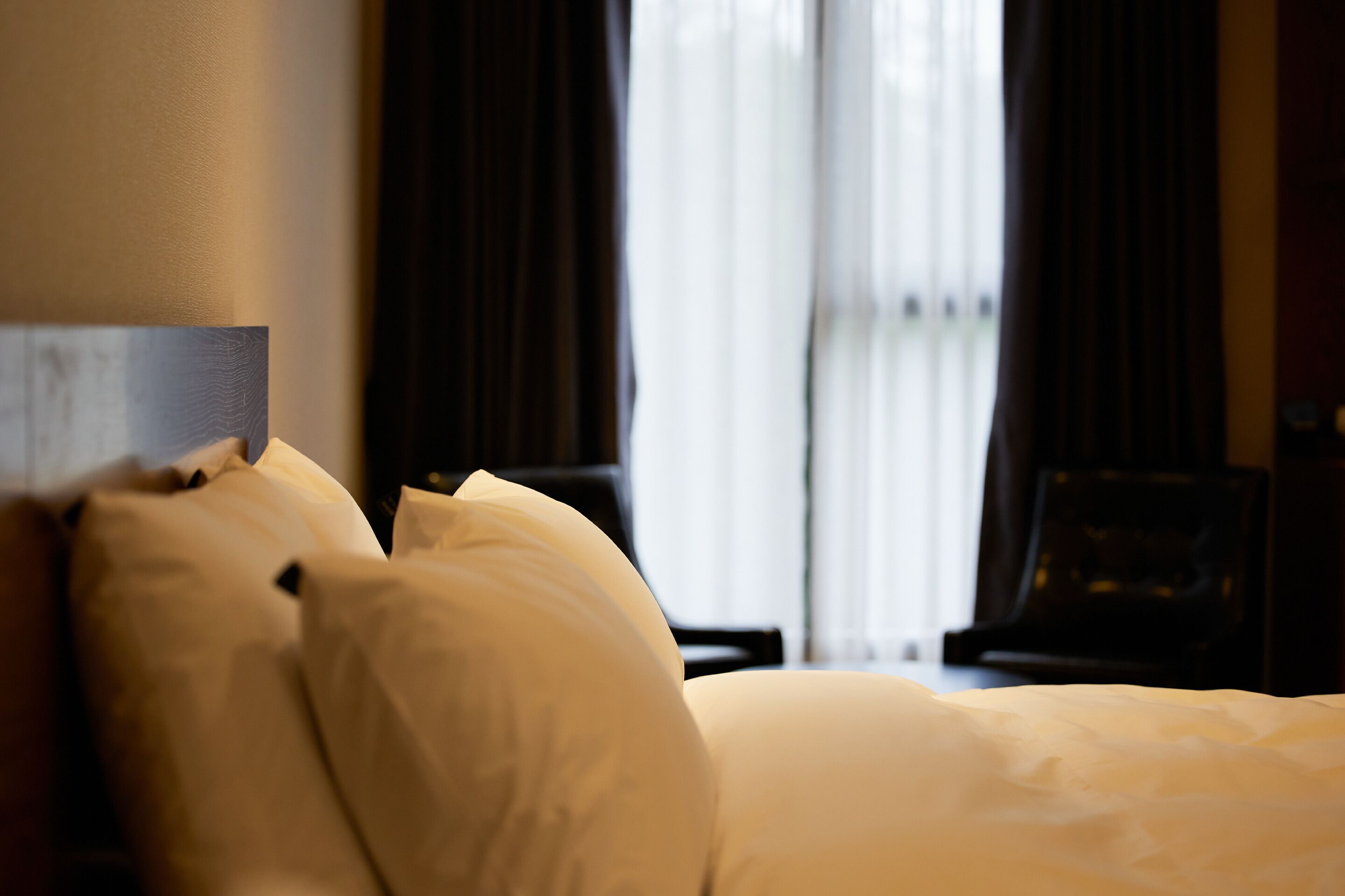 Een hotelkamer met een net opgemaakt bed, donkere gordijnen en een raam met natuurlijk licht.