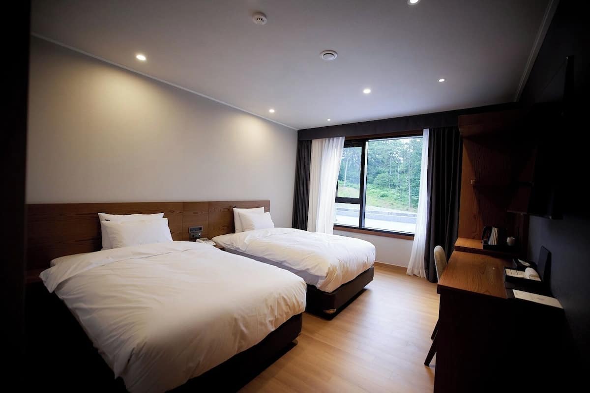 A hotel room with two beds, a desk, a chair, a window with a view of greenery, and a ceiling with recessed lighting.