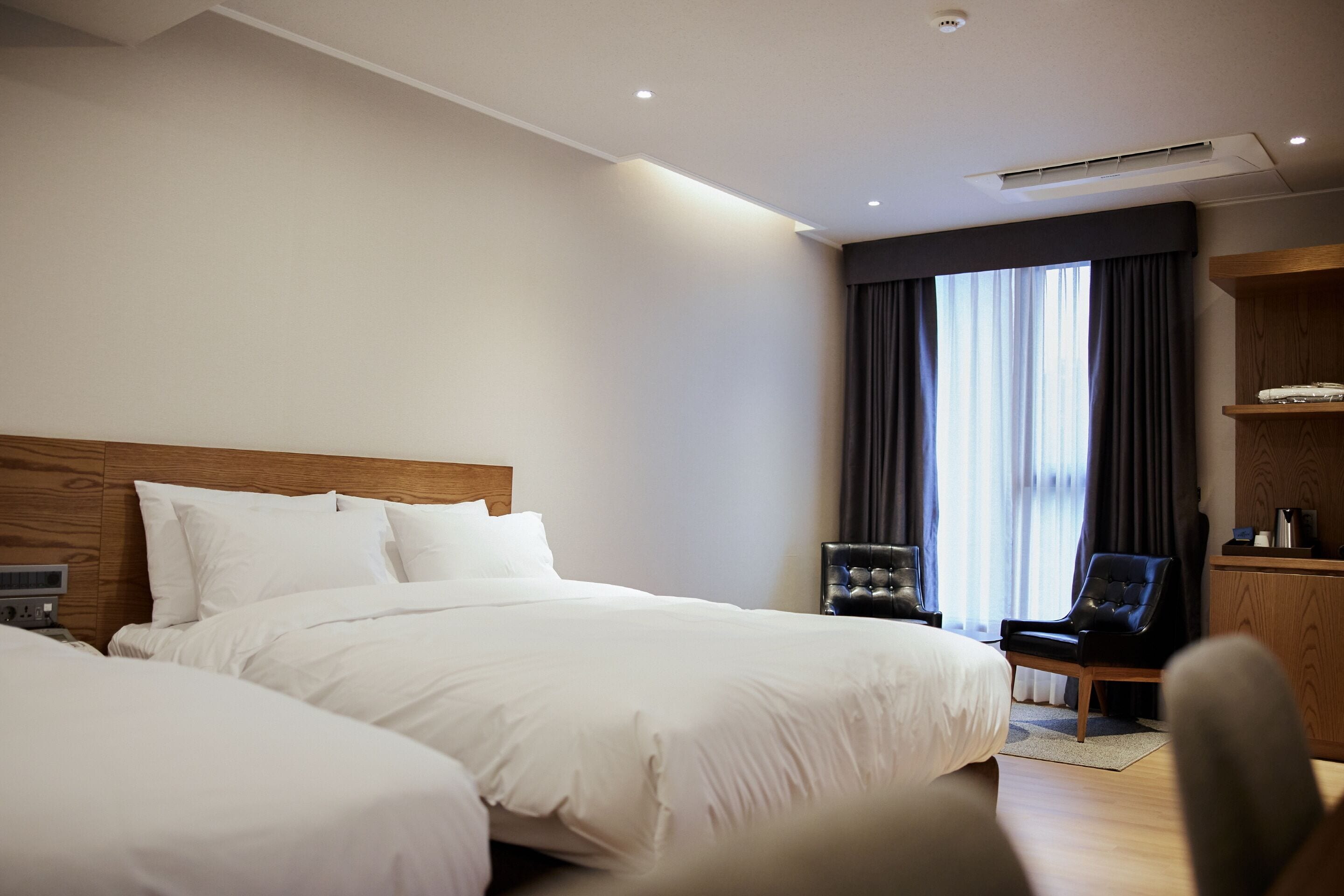 Quarto de hotel com duas camas, cabeceira de madeira, escrivaninha com cadeira e uma janela grande com cortinas.