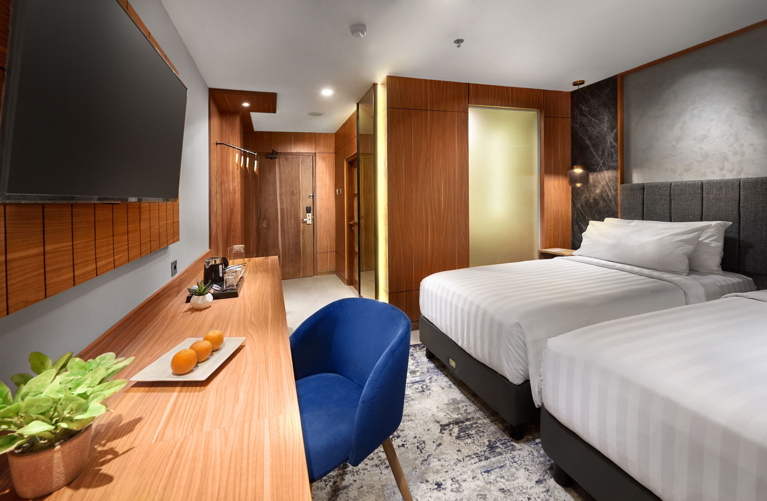 Une chambre d’hôtel moderne avec un bureau en bois, deux lits, une chaise bleue et un téléviseur.