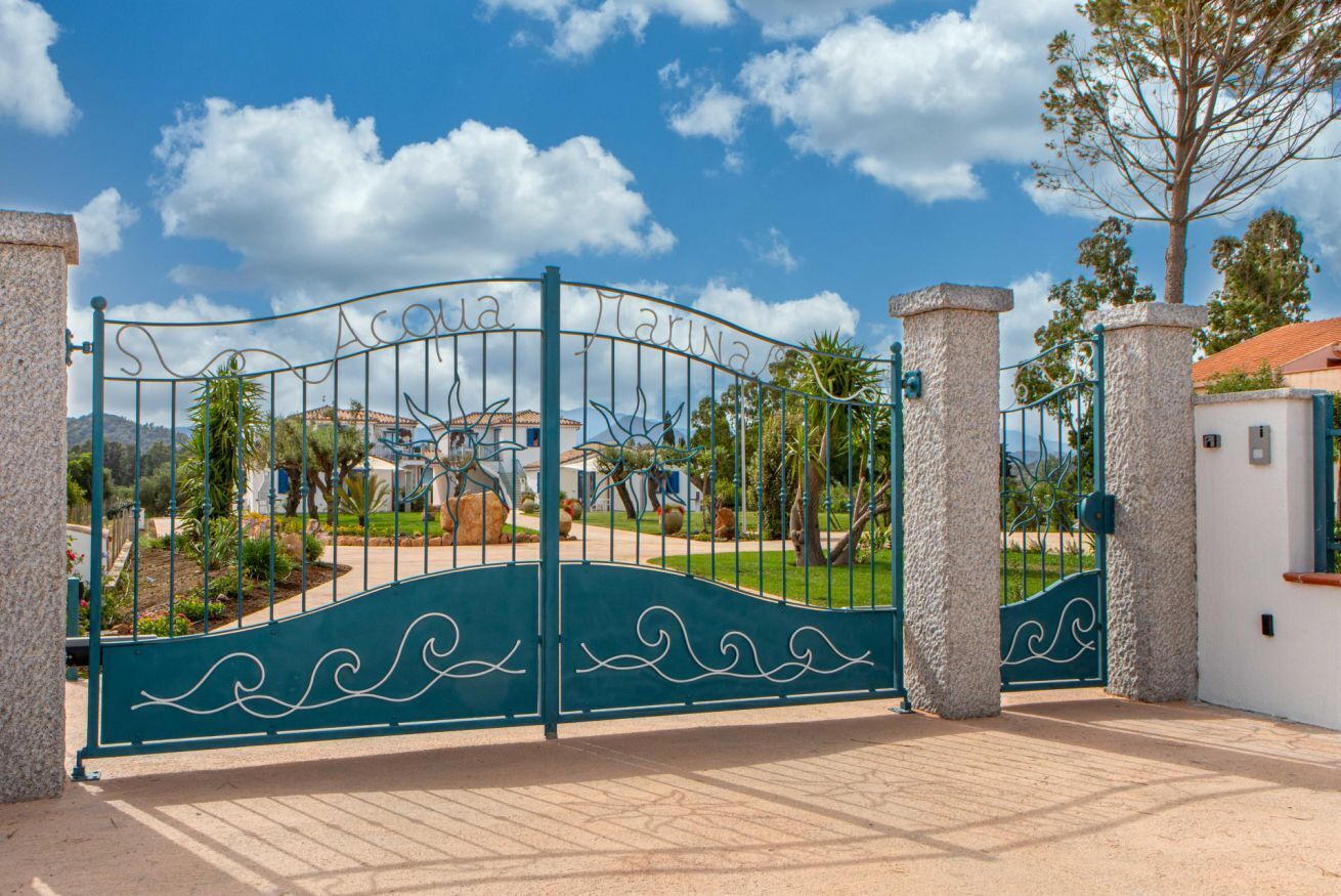 Una entrada protegida con una reja metálica decorativa y pilares de piedra, que conduce a un área ajardinada con árboles y un edificio al fondo.