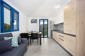 A modern kitchen with wooden cabinets, a dining area with a table and chairs, and a blue-framed window overlooking a scenic view.