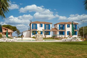 A two-story building with a pool, white walls, blue shutters, and a stone fountain.