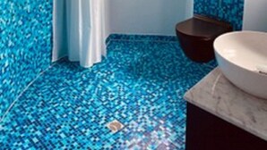A bathroom with a blue mosaic floor, a white bathtub, a dark wooden stool, and a window with a view of the outside.