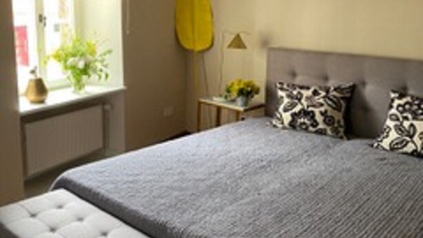 A bedroom with a large bed, a window with a yellow blind, and a bedside table with a lamp.