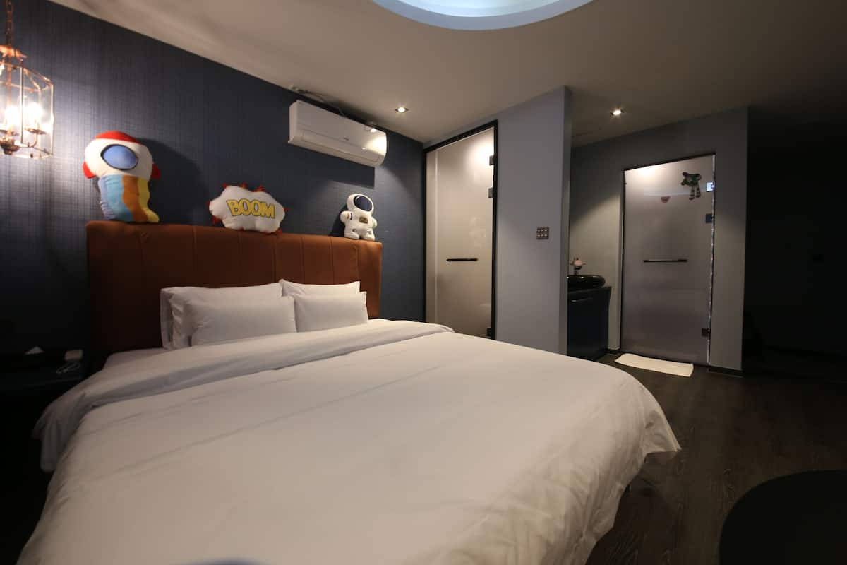 Una habitación de hotel moderna con una cama grande, cabecera con peluches, unidad de aire acondicionado y puerta que da a un baño.
