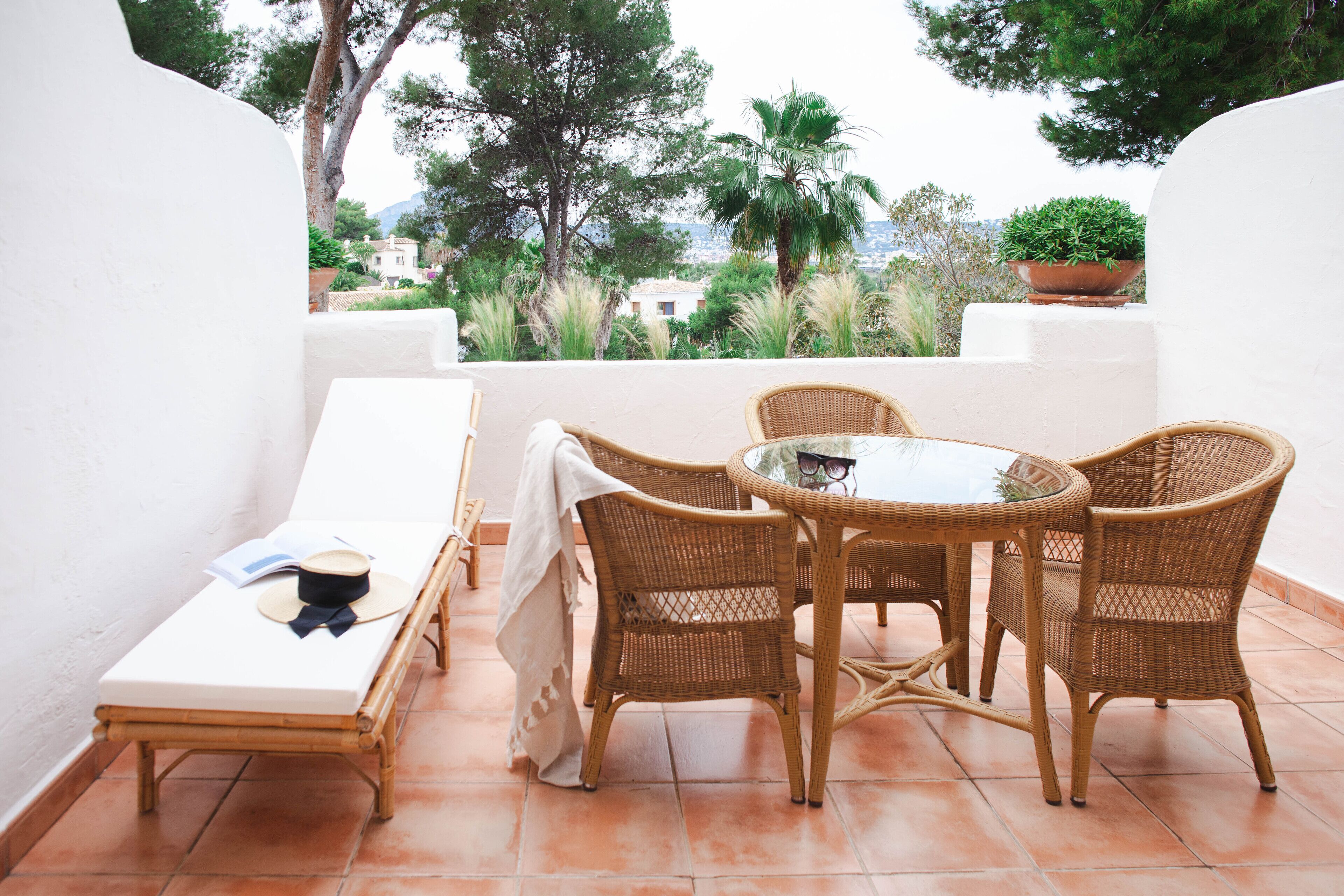 A terrace with a white wall, a sun lounger, a table with wicker chairs, and a potted plant.