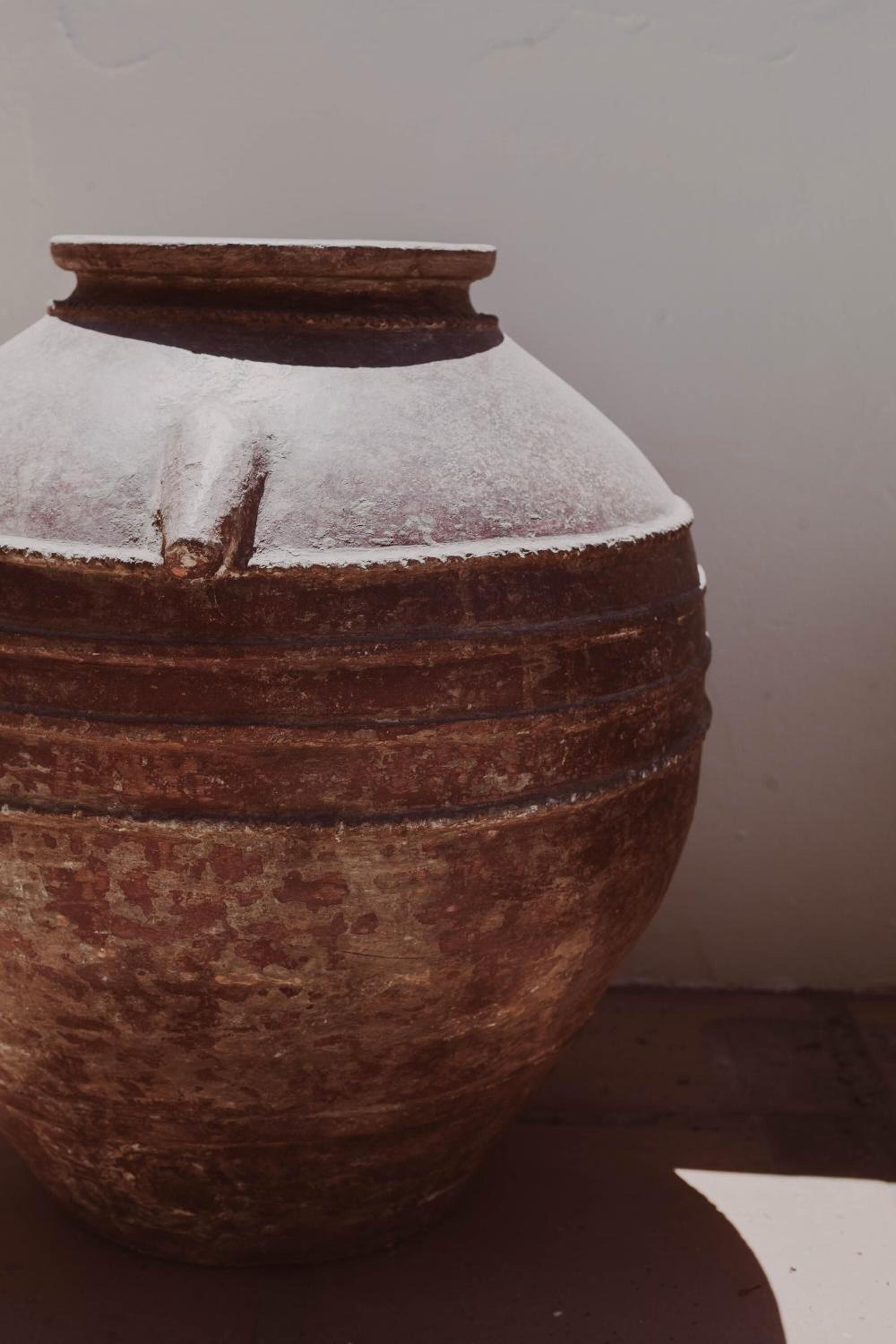 Un grand pot en terre cuite, de style rustique, avec une base large et un sommet étroit, posé sur une surface plane, adossé à un mur uni.