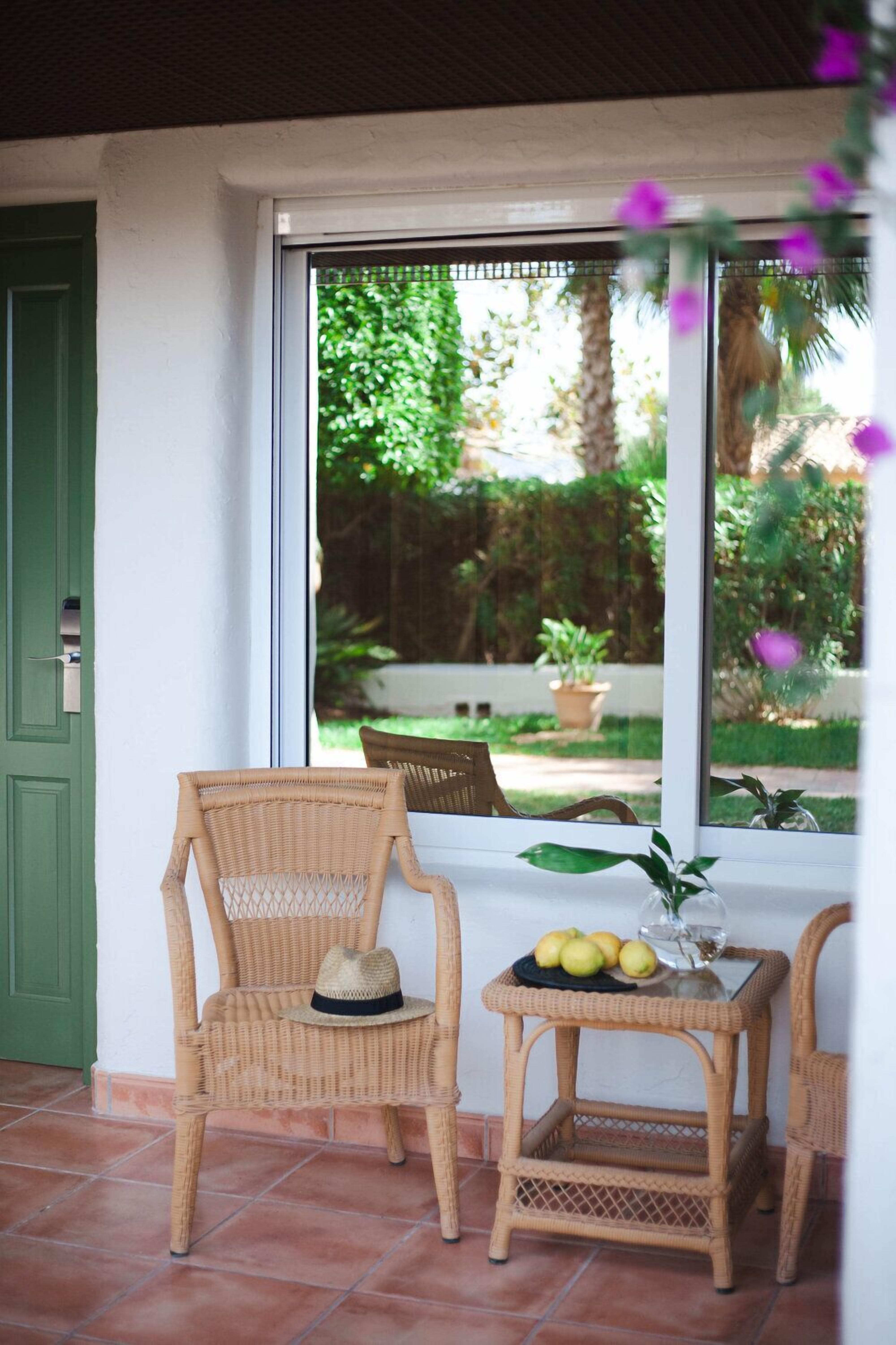 Une chaise et une table en osier, une assiette de fruits, une porte verte et, par la fenêtre, une vue sur un jardin.
