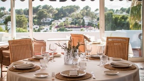 A round dining table set with plates, cutlery, and glasses, surrounded by wicker chairs, with a view of a residential area through large windows. - Ritual de Terra Javea Resort & Spa (Javea)