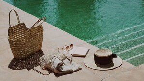 A poolside setting with a woven bag, a pair of sunglasses, a book, a hat, and a cup on a tiled surface. - Ritual de Terra Javea Resort & Spa (Javea)