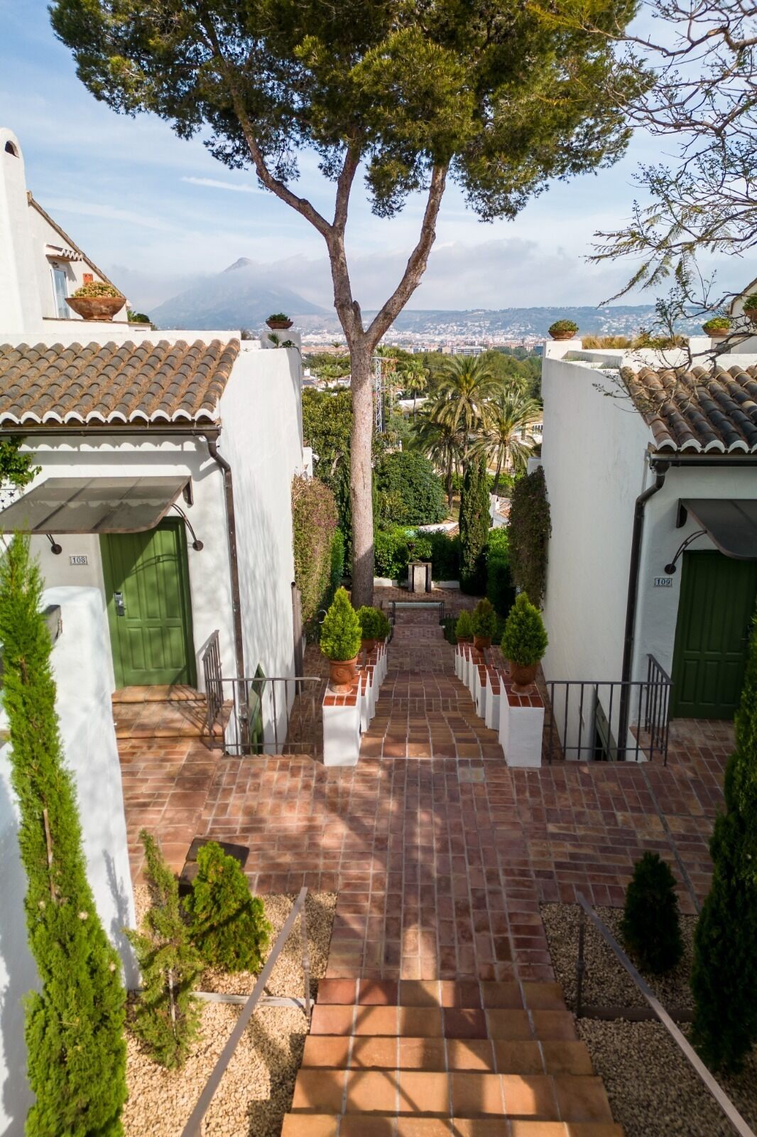 A courtyard with a central pathway flanked by white buildings with tiled roofs, surrounded by greenery and a view of distant mountains.