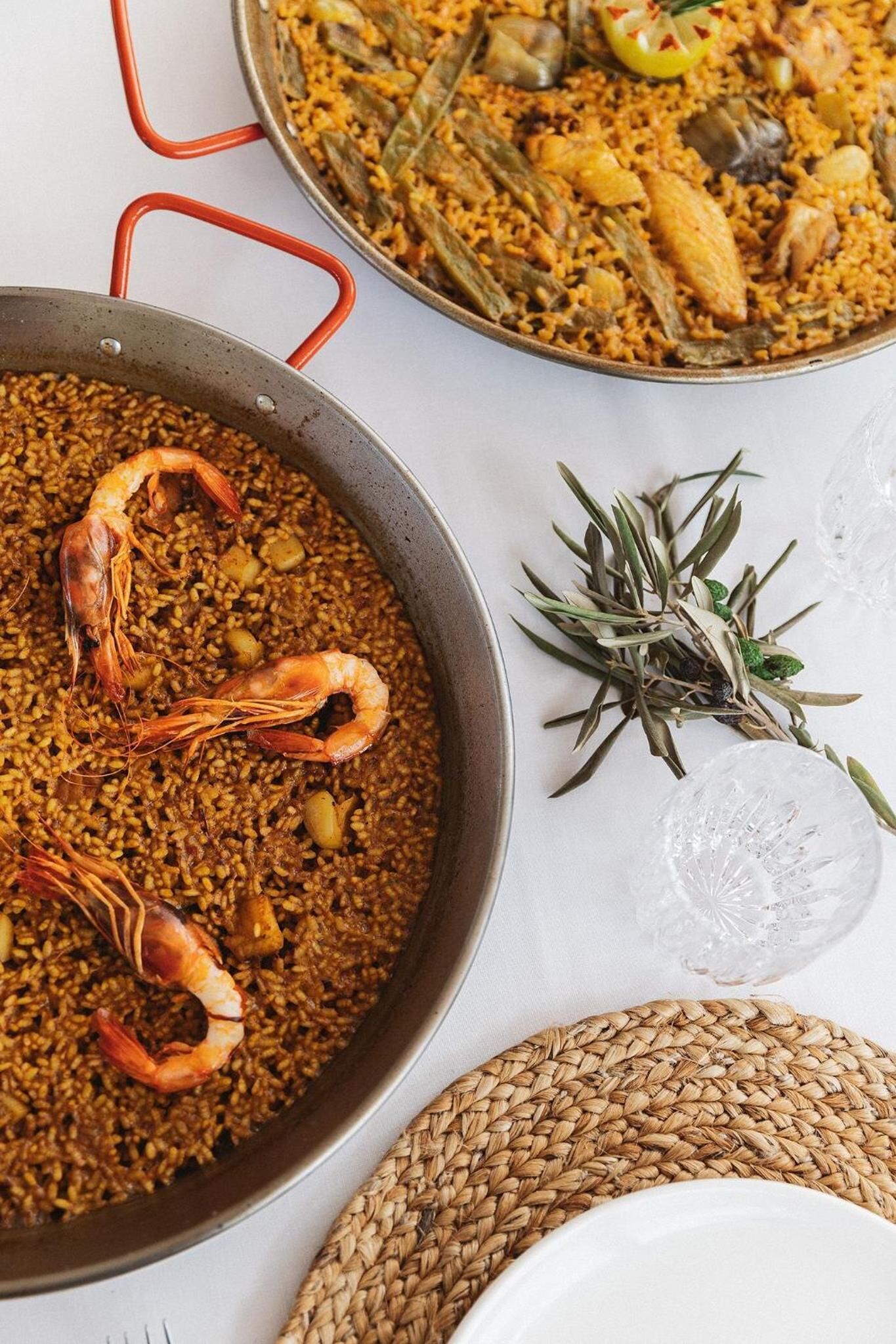 Een grote pan zeevruchtenpaella met garnalen, een kleinere pan met rijst en groenten, en een takje rozemarijn op een witte ondergrond.
