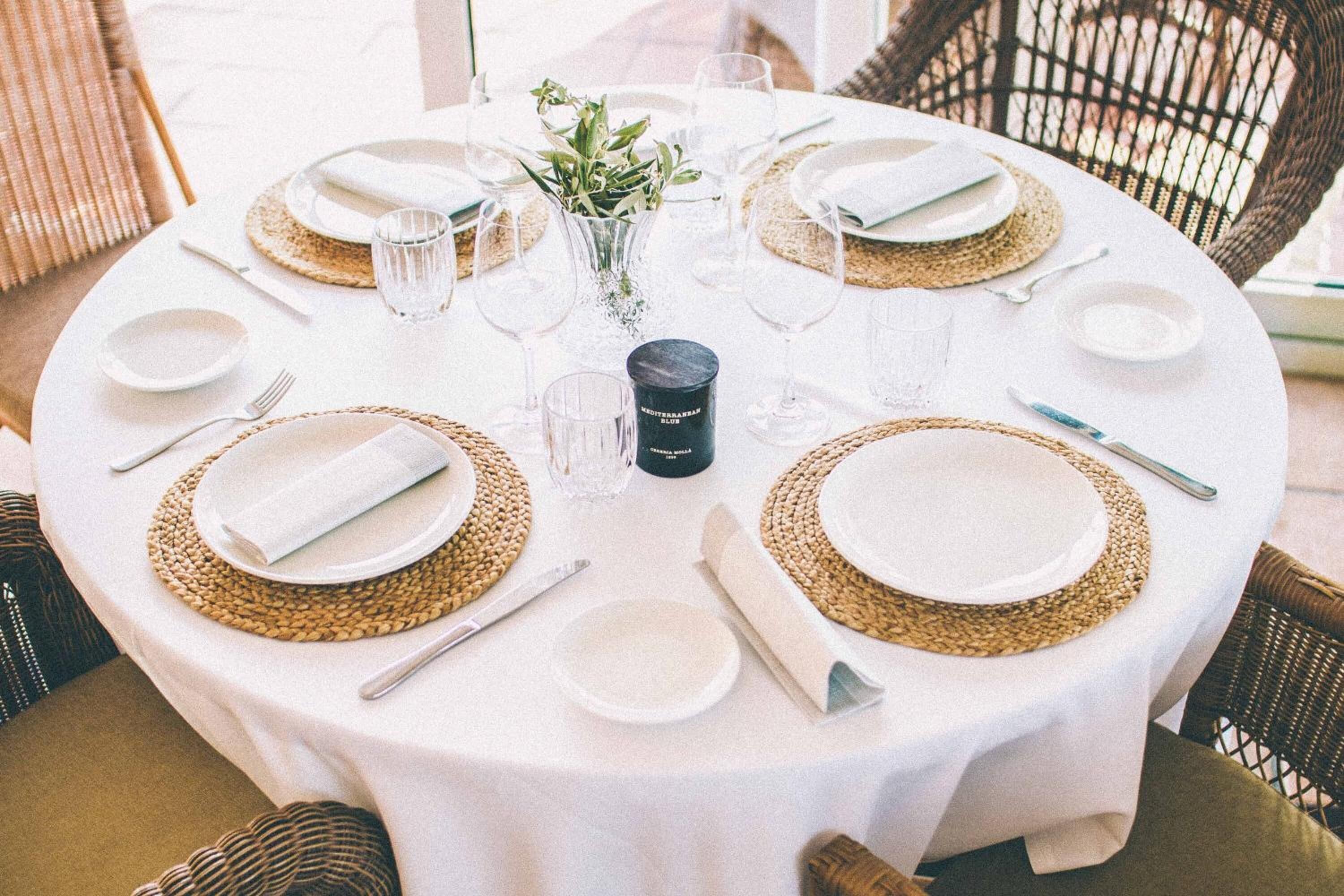 A round dining table with white tablecloth, woven placemats, and neatly arranged plates, glasses, and cutlery.