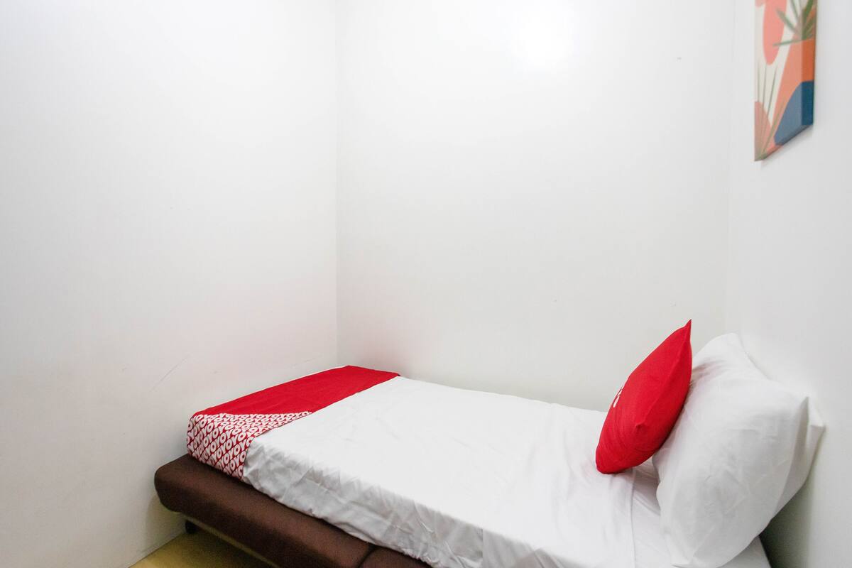 Une petite chambre avec un lit simple, recouverte d’une housse de couette blanche, agrémentée d’un oreiller rouge et d’une couverture à motifs rouges et blancs.