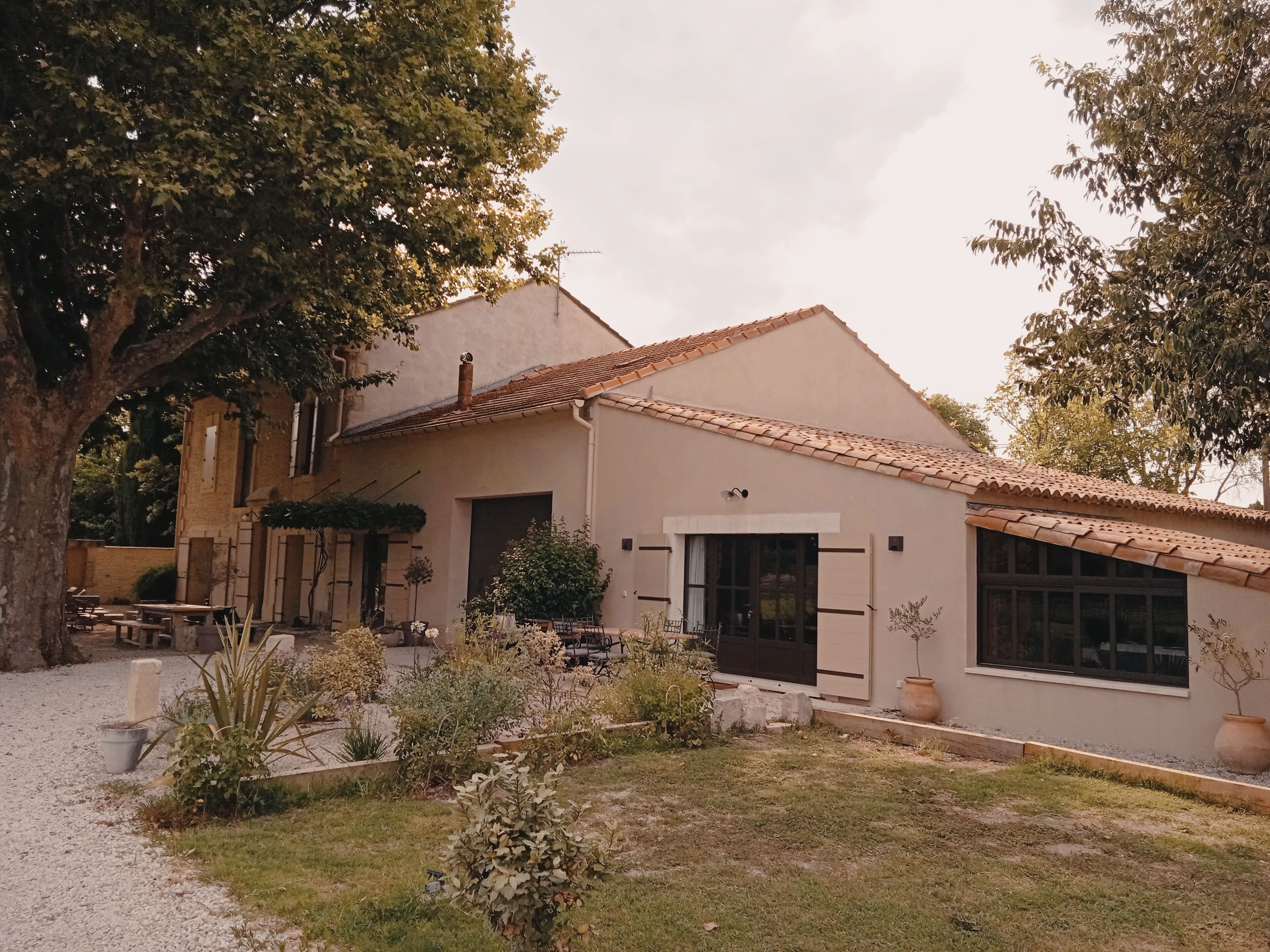 A two-story building with a tiled roof, a front door, and a small garden area with plants and a bench.