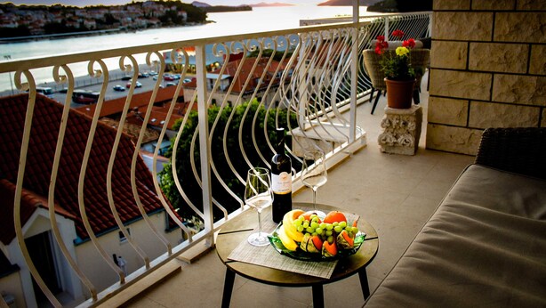 A balcony with a table set with fruit and wine glasses, overlooking a coastal town and a body of water. - Apartments Tayra (Dubrovnik)