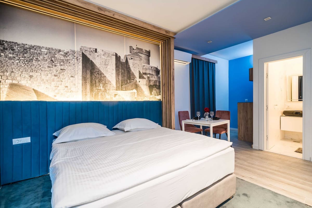 Une chambre d’hôtel avec un grand lit, un bureau avec une chaise, une petite table avec une chaise, et une salle de bain visible à travers une porte ouverte.