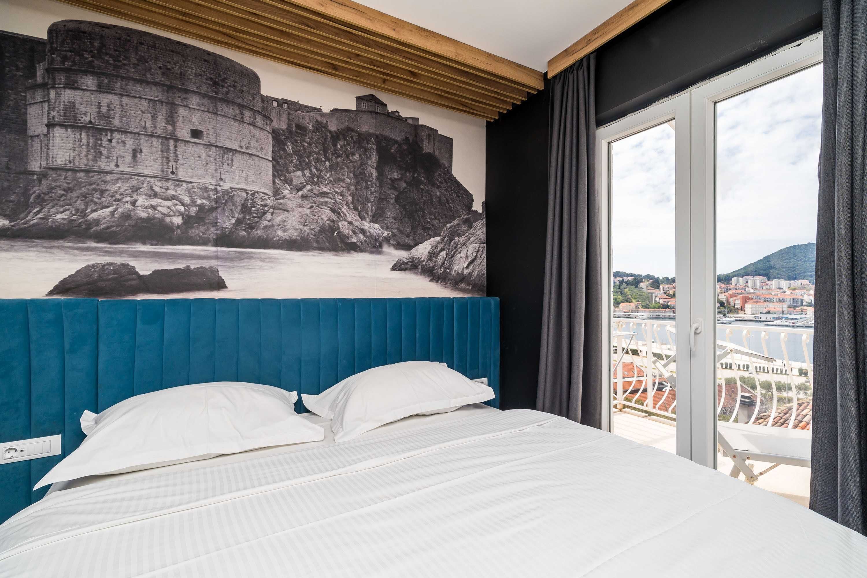 A hotel room with a bed, a view of a historic stone wall, and a balcony with a railing.