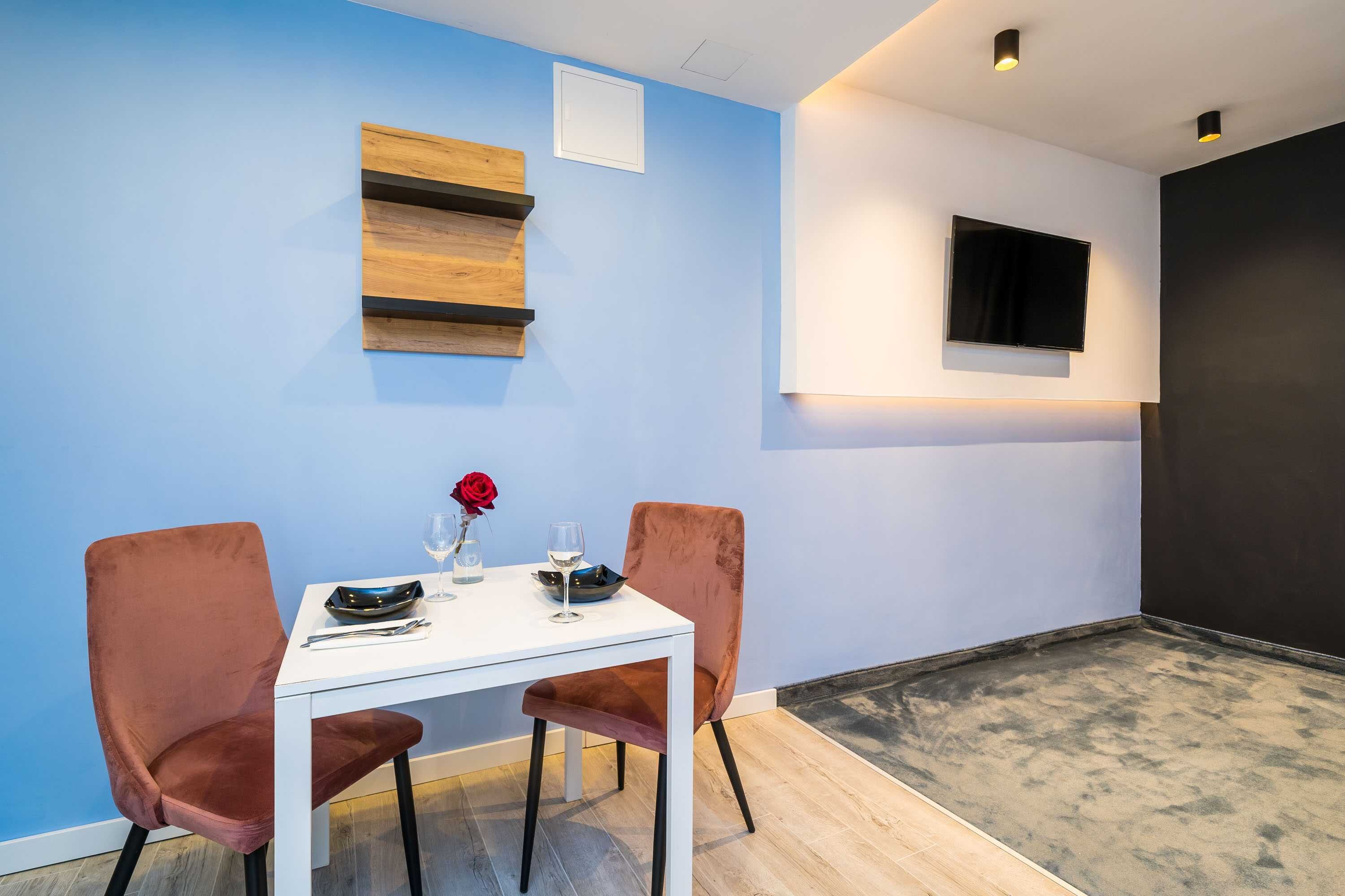 A dining area with a white table, two velvet chairs, a rose vase, and a flat-screen TV mounted on the wall.