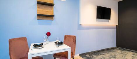 A dining area with a white table, two velvet chairs, a rose vase, and a flat-screen TV mounted on the wall.