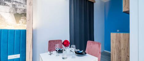 A dining area with a white table set for two, two pink chairs, a red rose in a glass vase, and a wooden cabinet in the background.