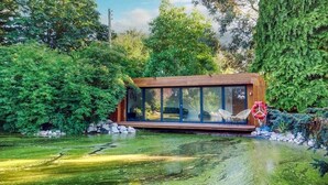 A modern cabin with large glass windows, situated by a pond surrounded by lush greenery.
