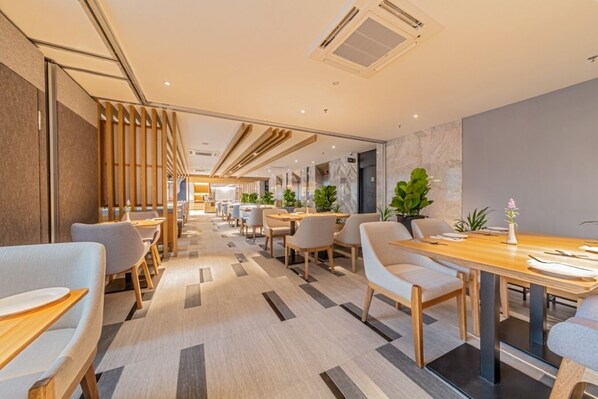 Un espace de restauration moderne dans un hôtel, avec de longues tables, des chaises et des plantes en pot.
