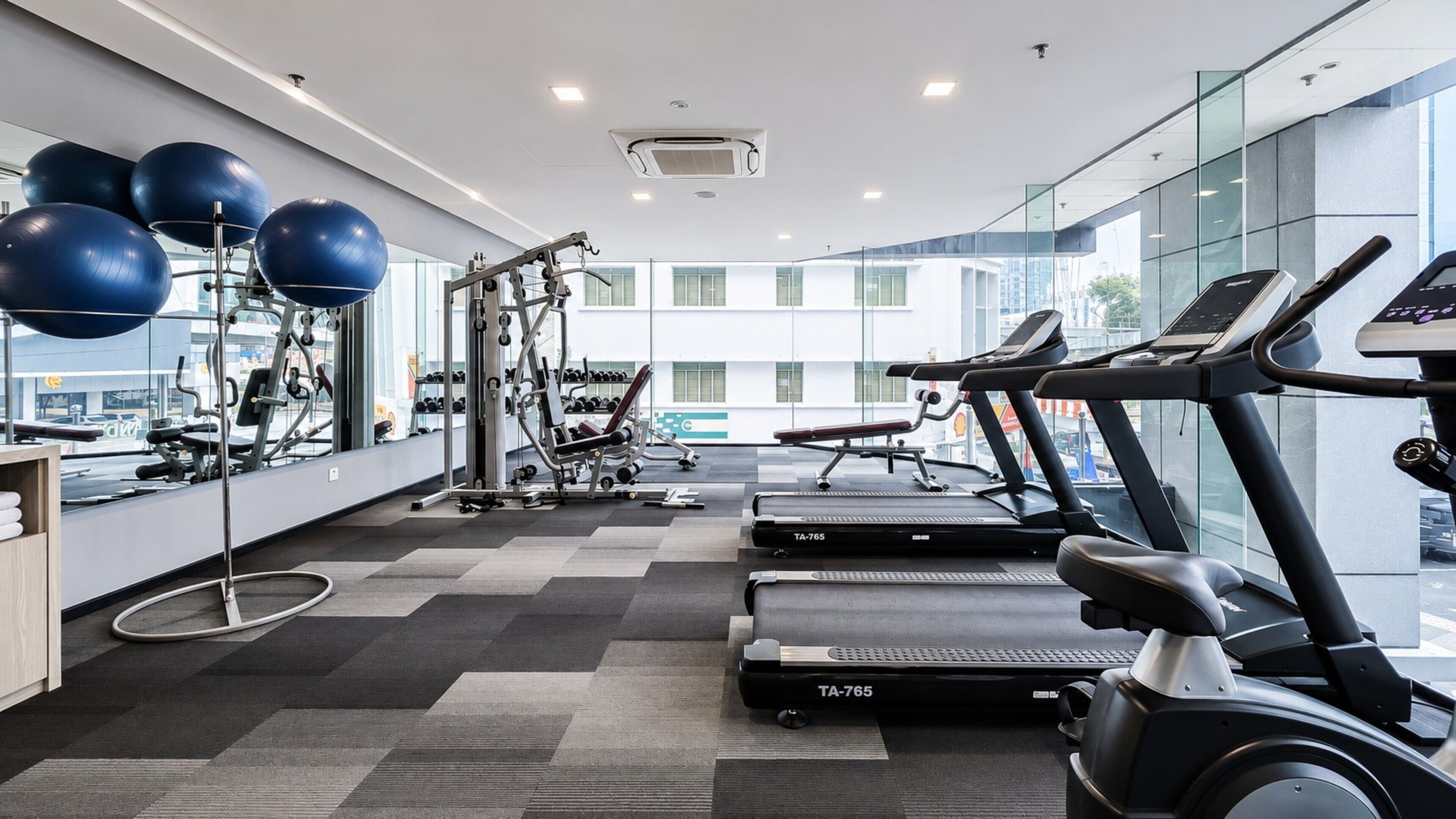 A modern gym with exercise equipment, including treadmills and a weight machine, and a large window offering a view of the city.