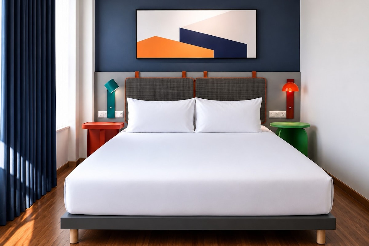 Ein modernes Hotelzimmer mit einem großen Bett, einem geometrisch gestalteten Kopfteil, einem Nachttisch mit Lampe und einem Wandbild.