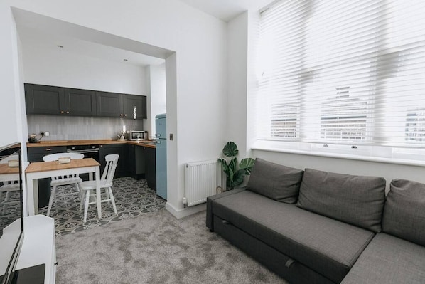 A compact living space with a kitchenette, a sofa, a dining table with chairs, and a large window with blinds.