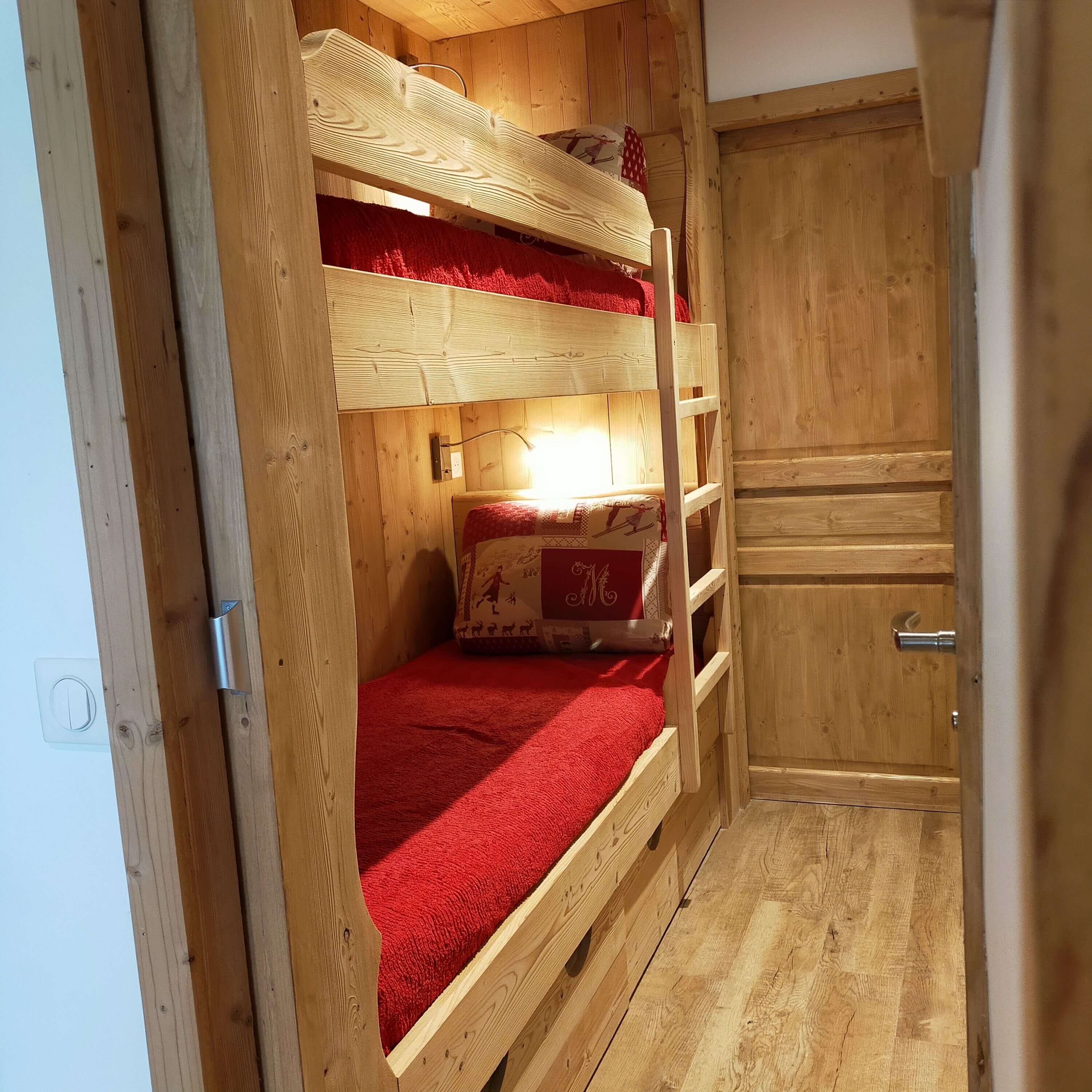 A wooden bunk bed room with a red blanket, a small table with a decorative pillow, and a wooden wardrobe.
