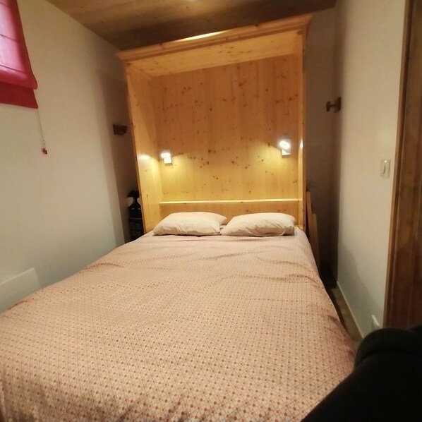 A narrow room with a bed, wooden headboard, and two wall-mounted lights. - Newly renovated apartment. It is located 150 meters from the slopes, across from the community center (Montvalezan)