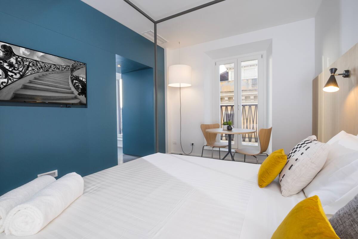 A hotel room with a bed, a desk, and a chair. There is a window with a view of buildings outside.
