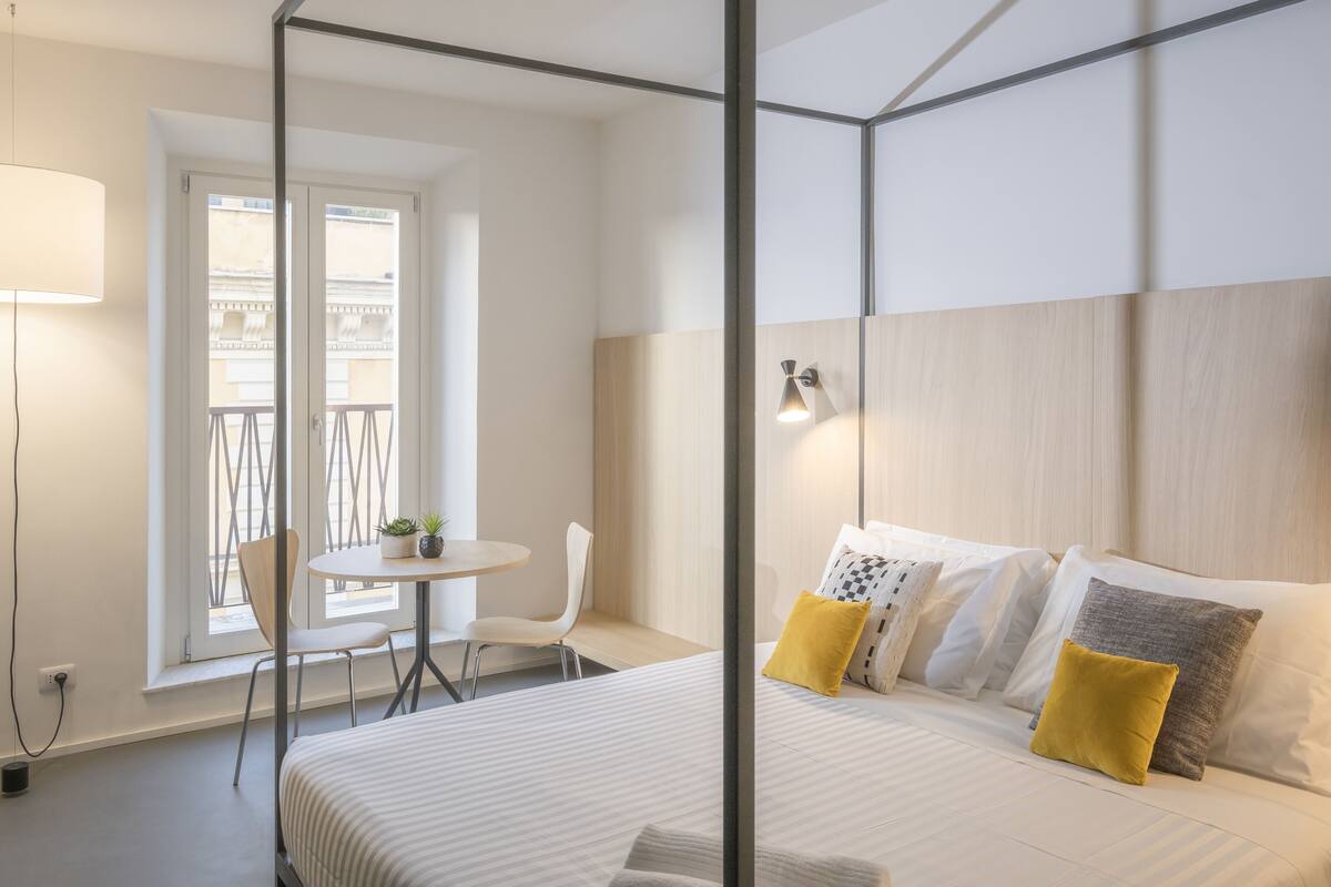 A modern hotel room with a four-poster bed, a small round table with chairs, a balcony with a view, and a wall-mounted lamp.