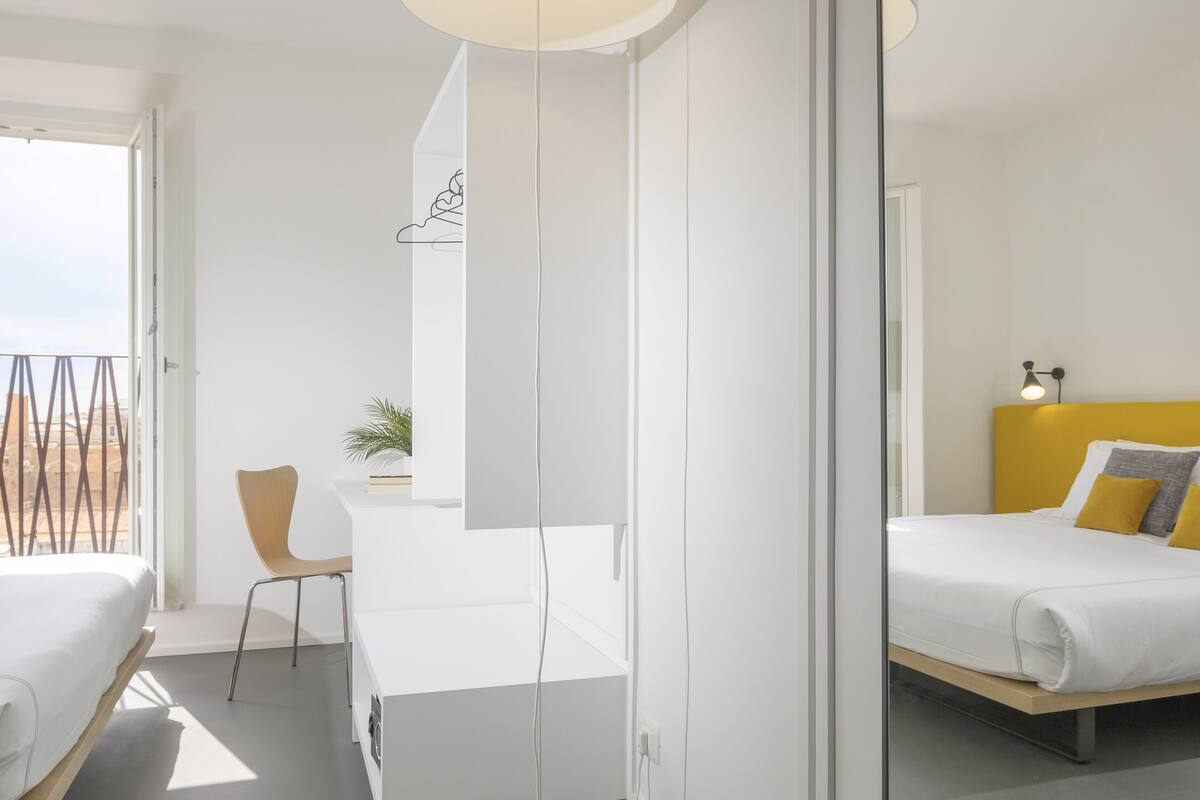 A modern hotel room with a balcony, a desk, and a bed with yellow and gray bedding.
