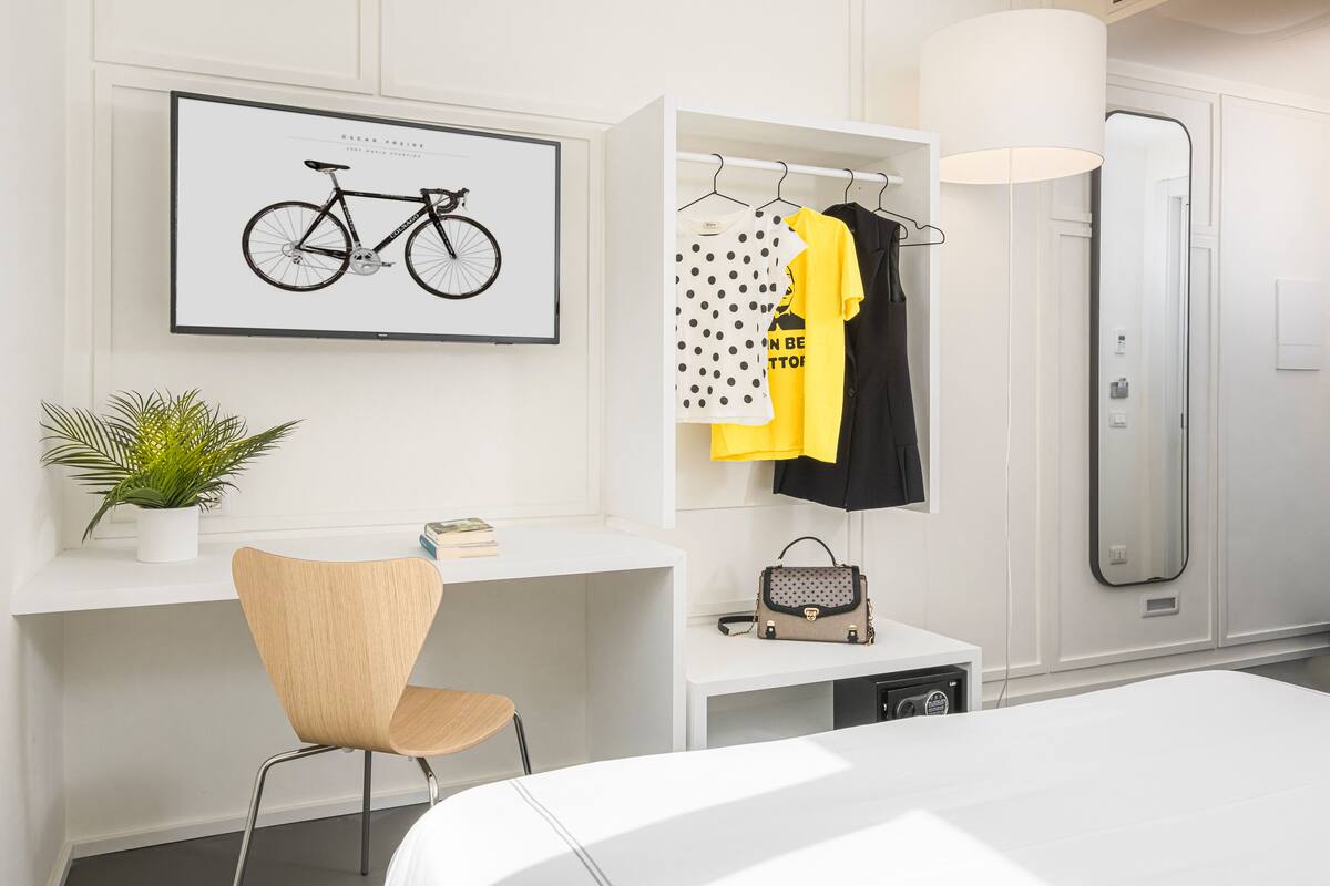 A modern hotel room with a white desk, a chair, a framed bicycle picture, a clothing rack, and a large mirror.