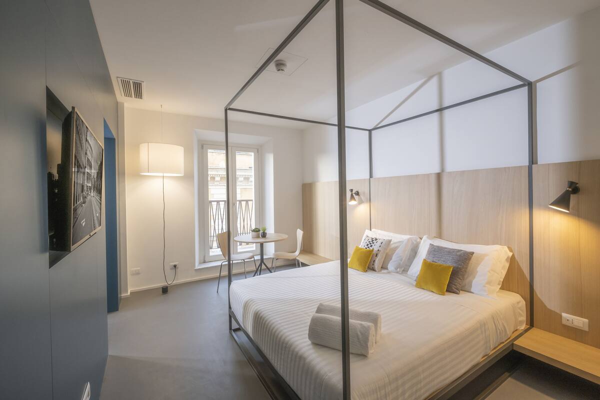 A modern bedroom with a four-poster bed, a small table with chairs, a lamp, and a view of the outdoors.