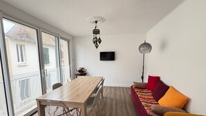 A modern apartment with a wooden dining table, a sofa with colorful cushions, a TV mounted on the wall, and a large window offering a view of the outside. - Charming City Center House with Beach Access, Free WiFi, Garage, Baby Gear, & Renovated Interiors (Les Sables-d'Olonne)