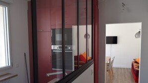 A modern kitchen with red cabinets, a glass door, and a dining area with a red sofa and a TV on the wall. - Charming City Center House with Beach Access, Free WiFi, Garage, Baby Gear, & Renovated Interiors (Les Sables-d'Olonne)