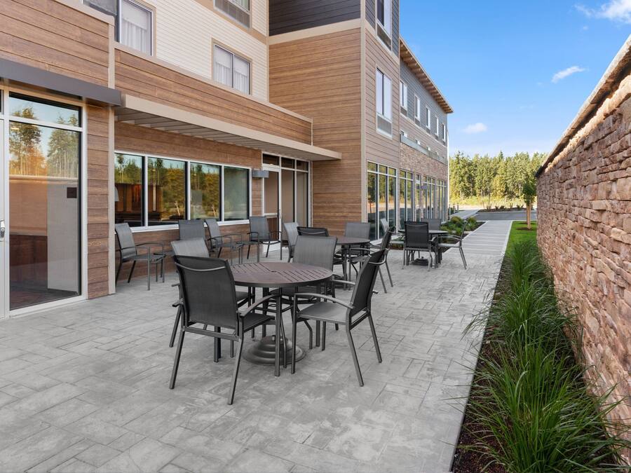 An outdoor patio area with multiple tables and chairs, a brick wall on one side, and a building with large windows and a wooden facade.