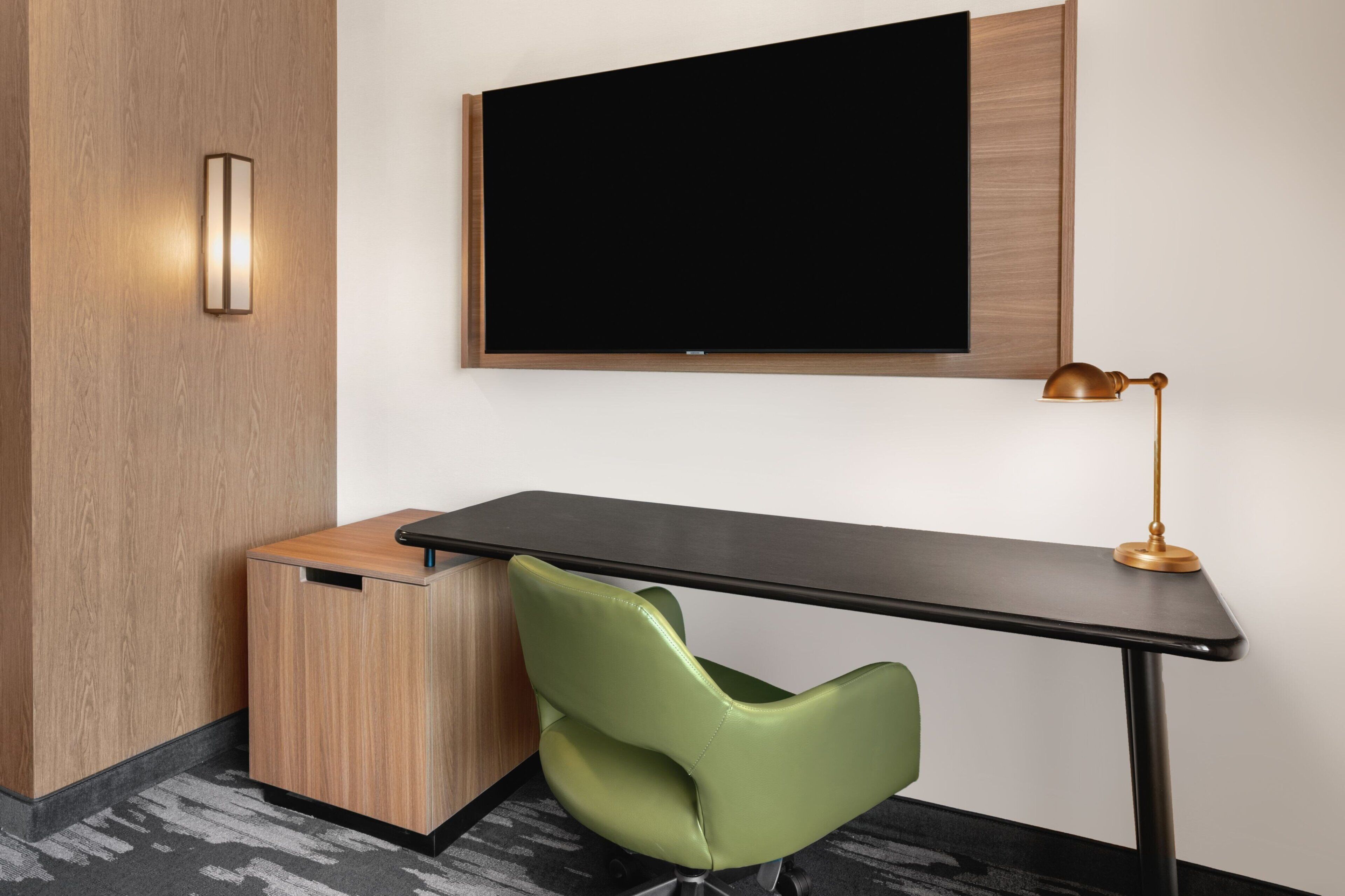 A modern room with a desk, a green chair, a television mounted on the wall, and a small cabinet.