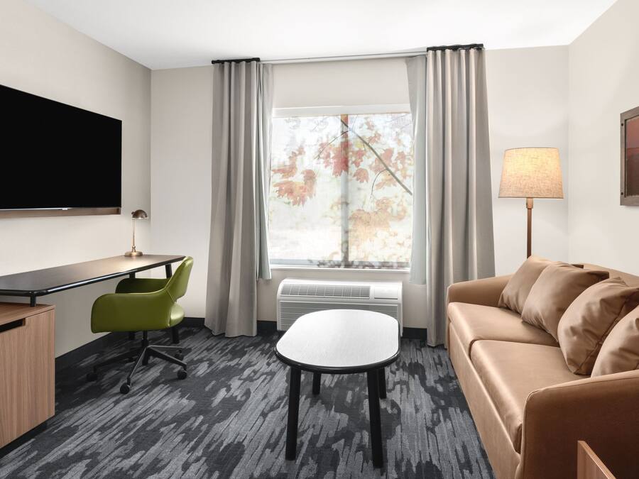 A hotel room with a flat-screen TV, a desk, a green chair, a brown sofa, a small table, and a window with curtains.