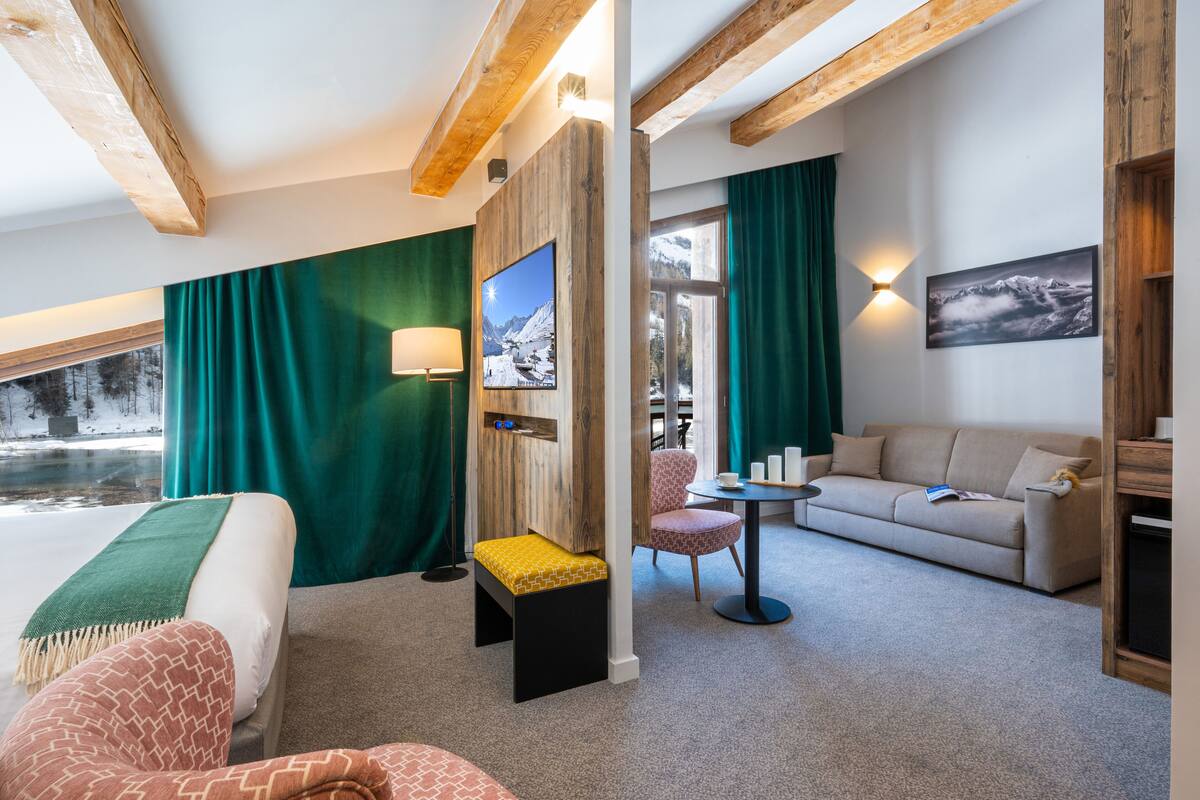 Habitación de hotel con cama, sofá, una mesita, una silla y vista a un paisaje nevado a través de una ventana.