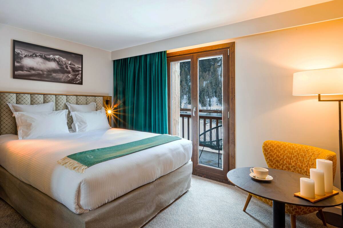 Habitación de hotel con cama, una mesa con velas, una silla, un balcón con vistas a árboles y un cuadro en la pared.