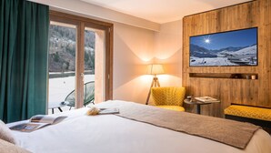 Ein Hotelzimmer mit Bett, Wandfernseher, Schreibtisch mit Lampe und Blick auf eine verschneite Landschaft durch das Fenster.