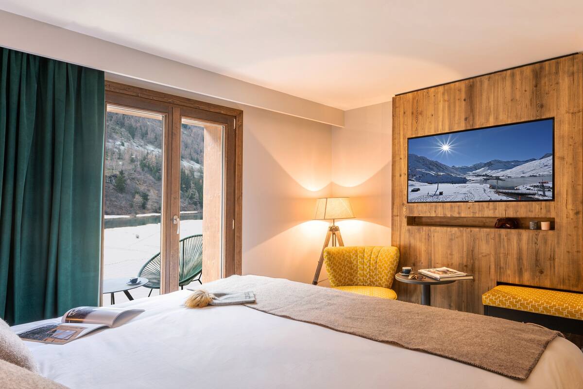 Una habitación de hotel con una cama, un televisor en la pared, un escritorio con lámpara y vistas a un paisaje nevado a través de la ventana.