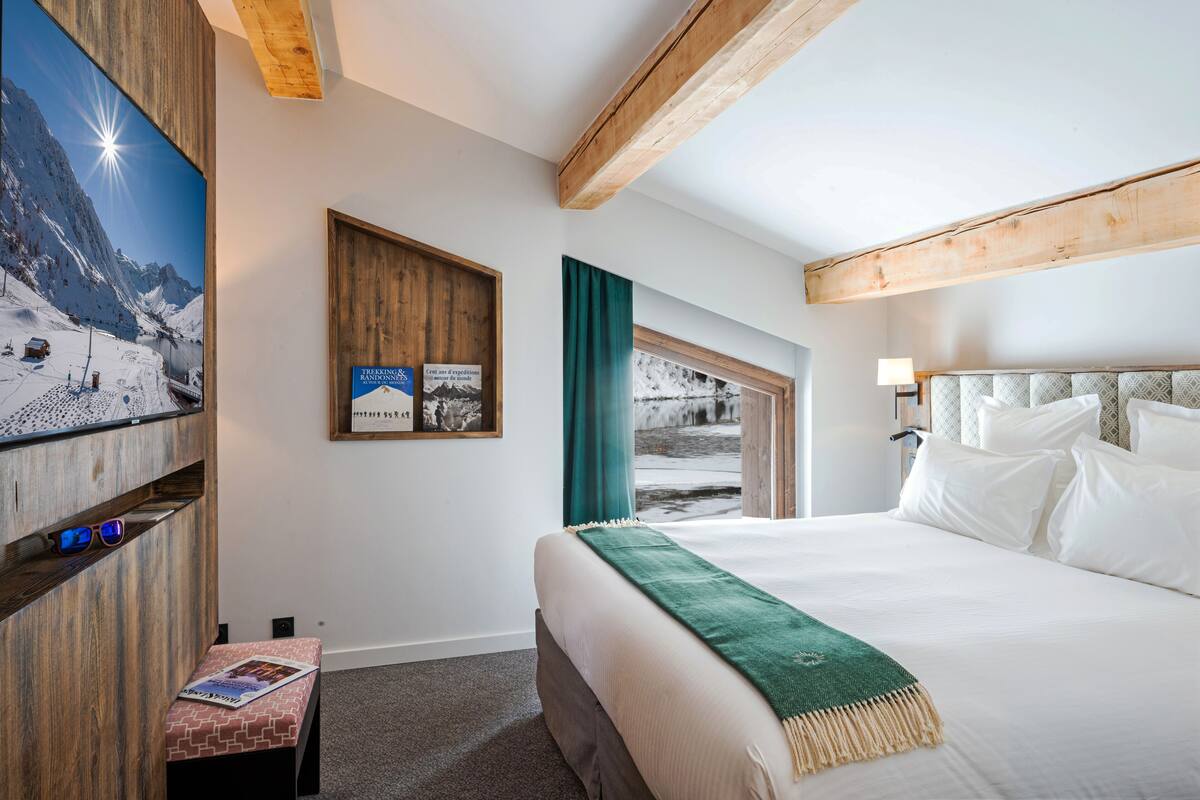 Una habitación de hotel con una cama, un televisor montado en la pared, techo de madera y vistas a un paisaje nevado a través de una ventana.